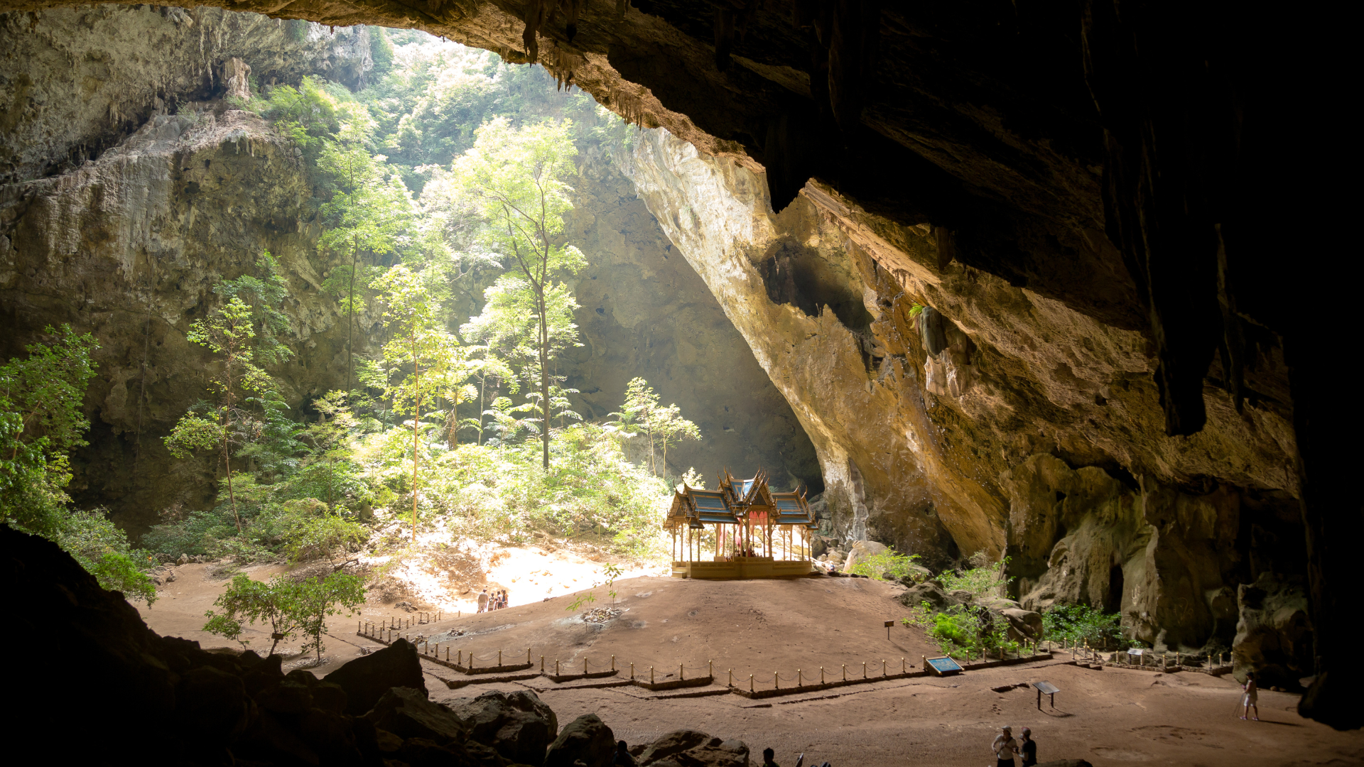 Photo of the inside of the Phraya Nakhon cave in Thailand. There is a small building on a hill inside the cave, trees in the background. 