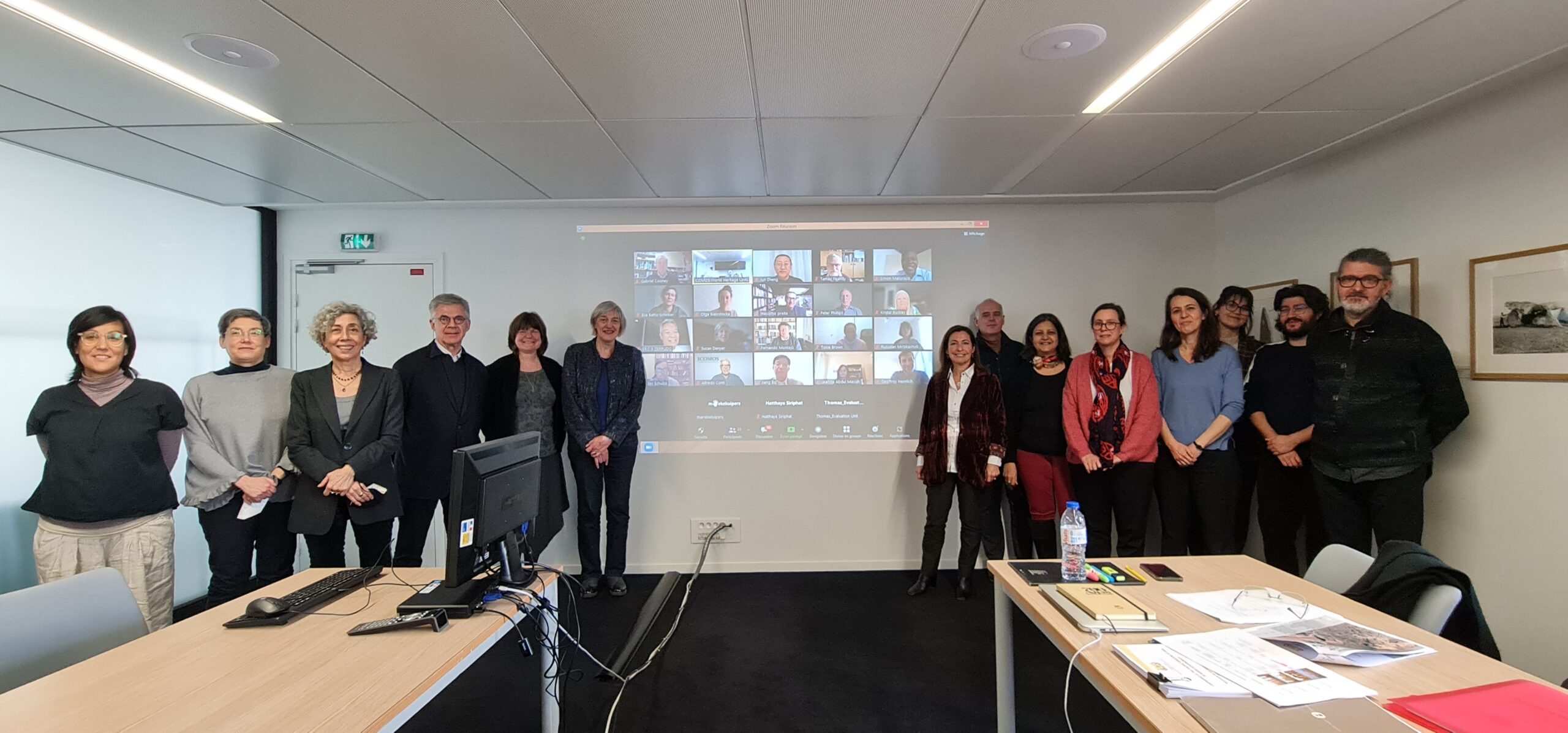 Photo de groupe. 14 personnes sont debout devant un mur sur lequel est projetée une session Zoom.