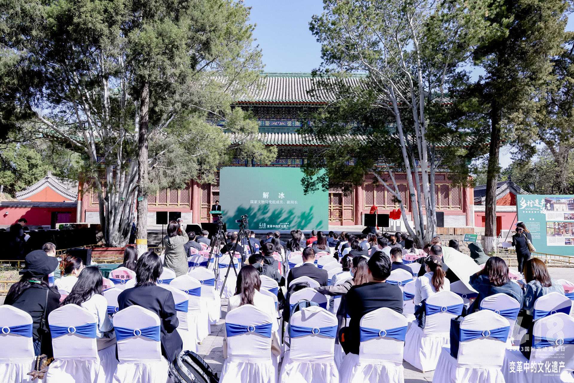 People sitting outdoors in front of a stage where a man is giving a speech