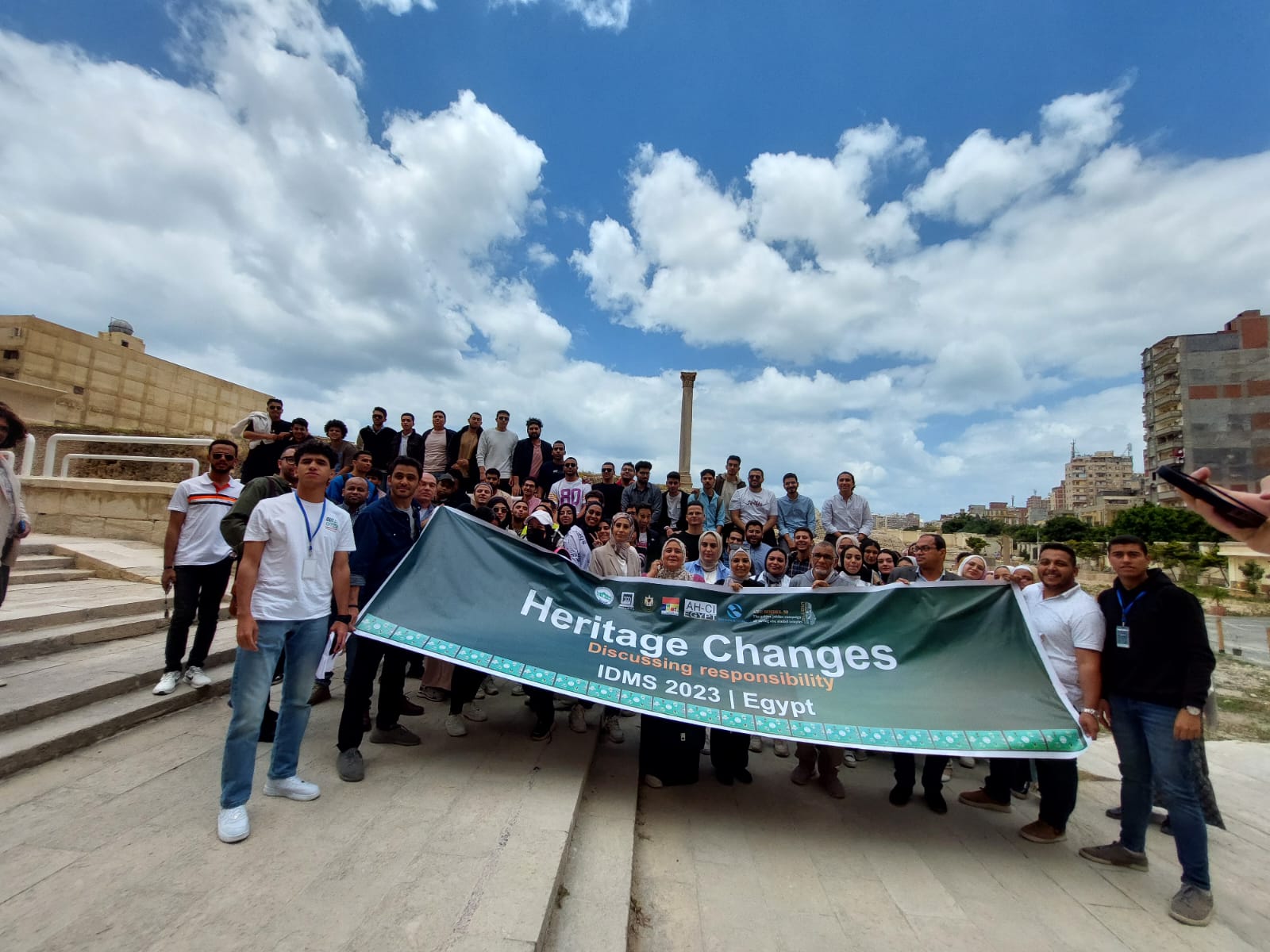 Group of around 50 people standing and holding a banner with "heritage changes, discussing responsibility, IDMS 2023 Egypt" written on it.