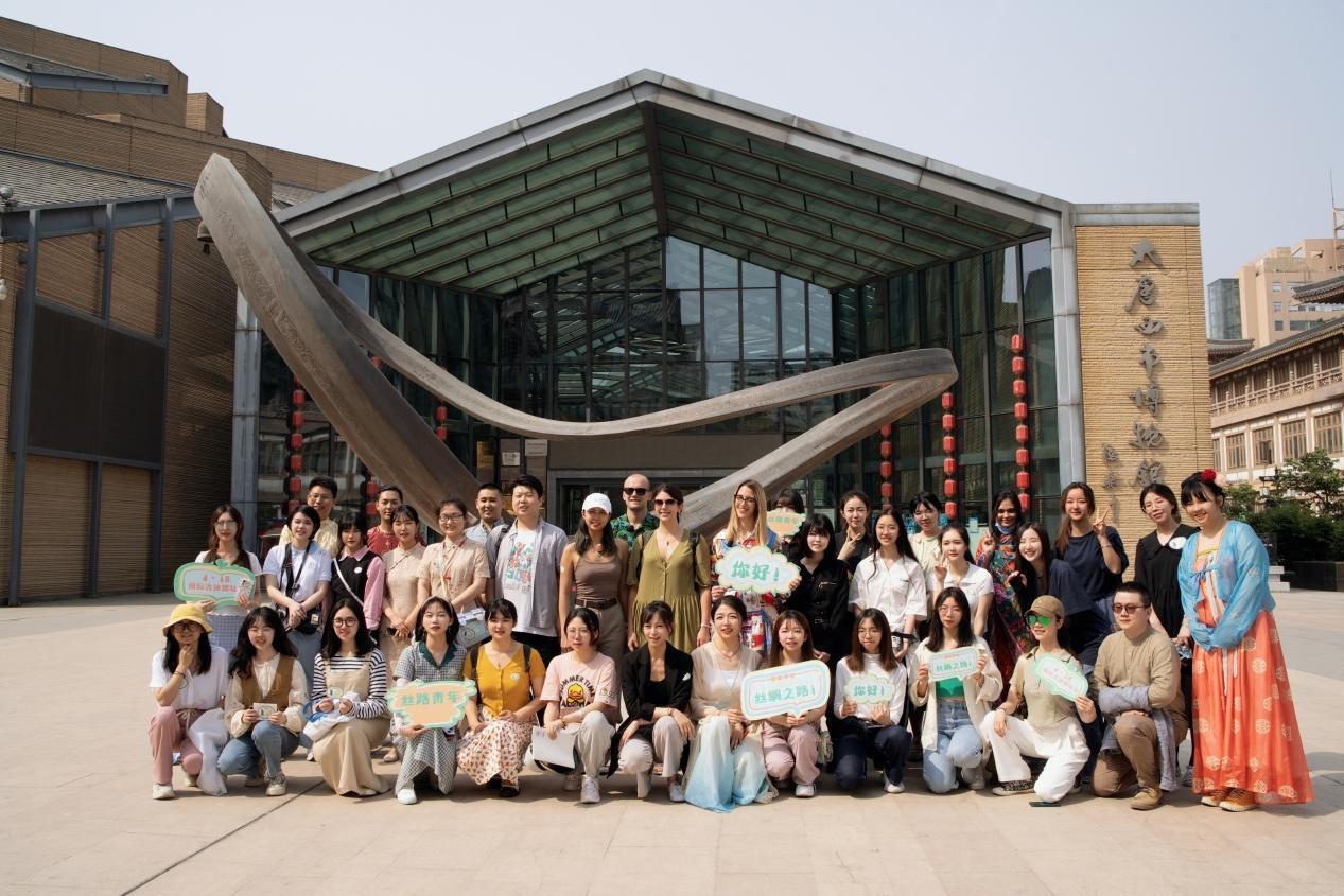 Group of 37 people smiling at the camera, standing or crouching, some of them are holding signs. They are standing in front of a building.