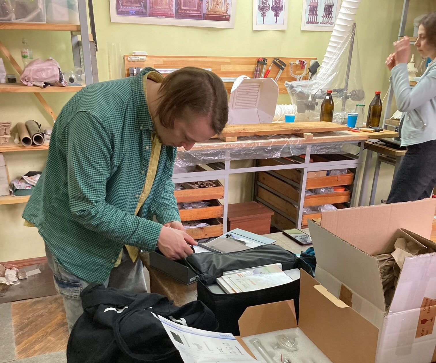 Man seen from the side, looking down at the content of a small black box laid on a table. On the table are various objects including cardboard boxes and sheets of paper. In the background, a woman is standing in front of a wooden workbench.