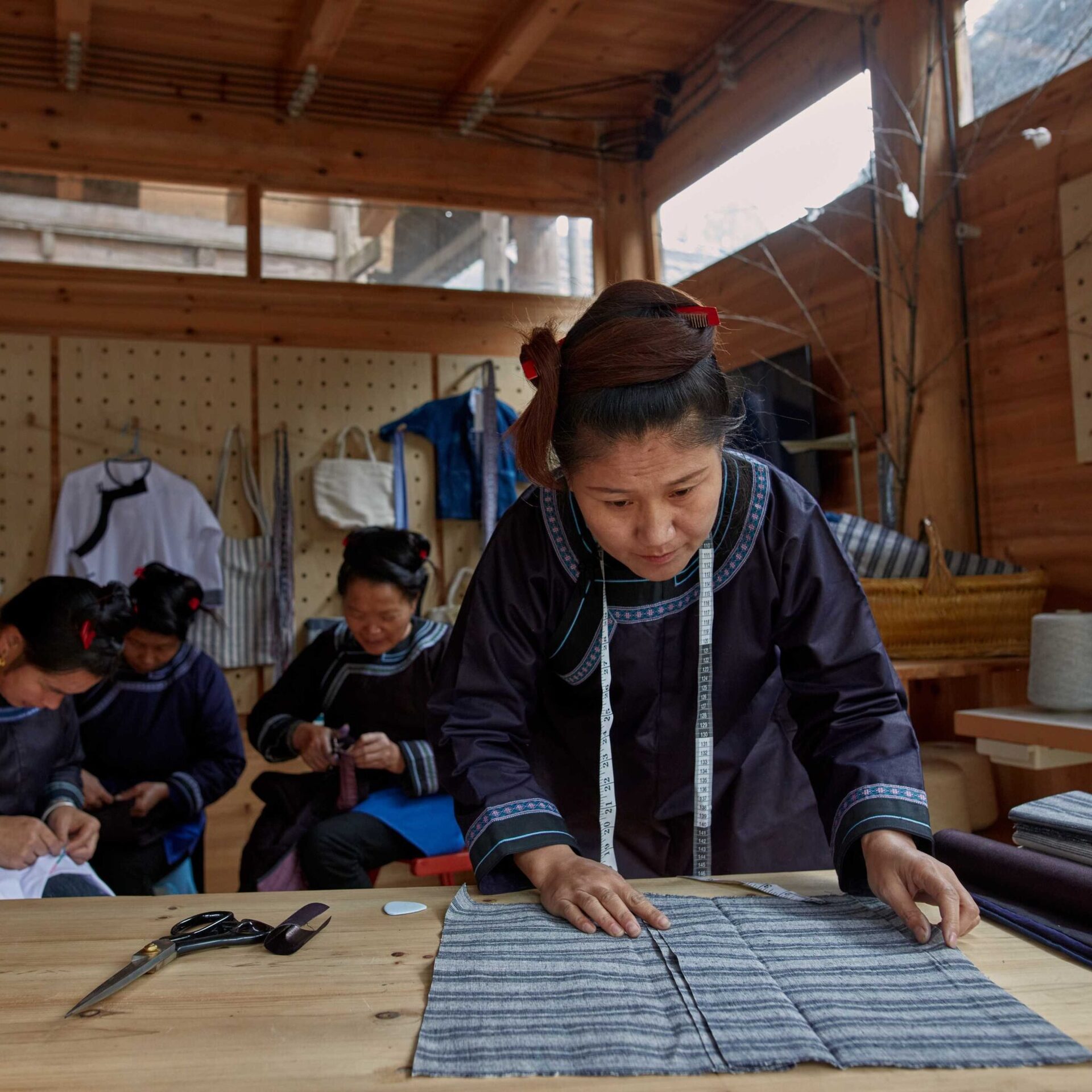 In the foreground, woman aligning pieces of fabric on a wooden table. In the background are 3 other women sitting, cutting and sewing fabric. 