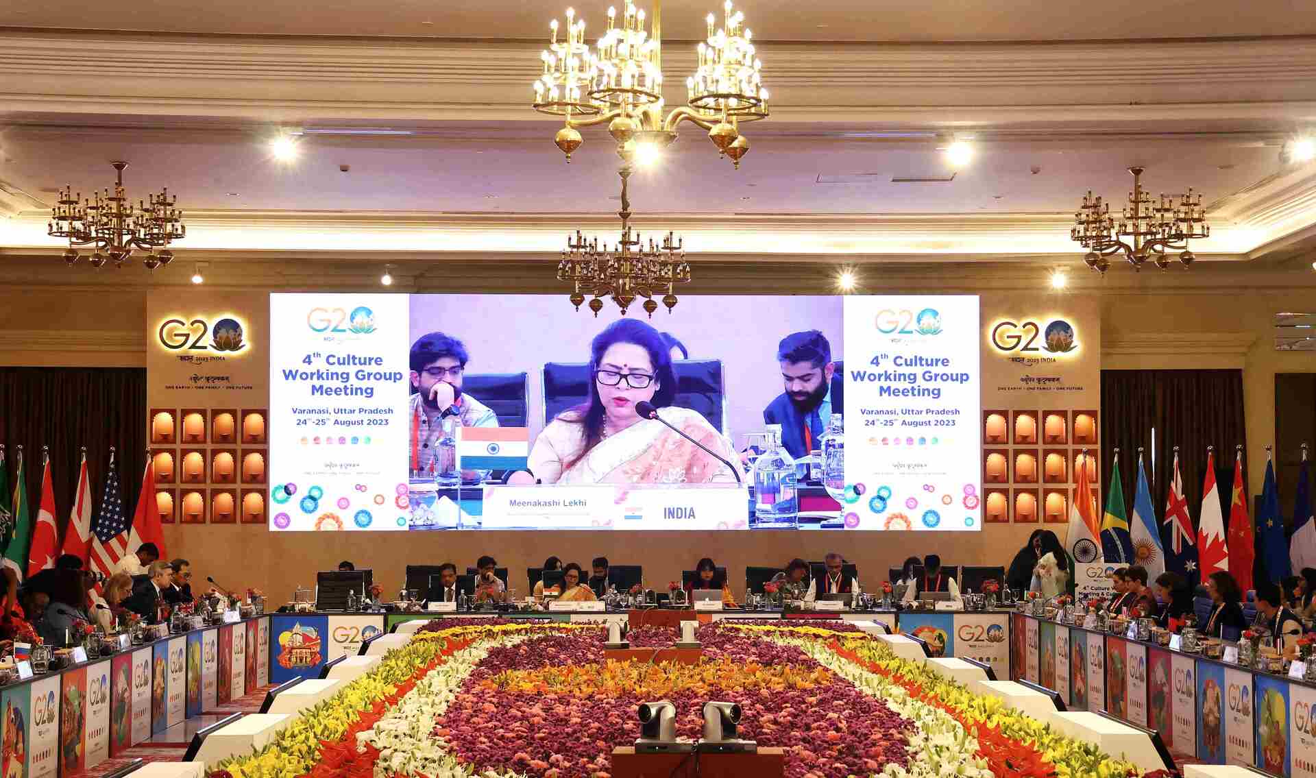 People sitting around an oval table. In the center of the room, there is an arrangement of flowers. The wall at the back of the room carries a screen, displaying a woman speaking in a microphone. 