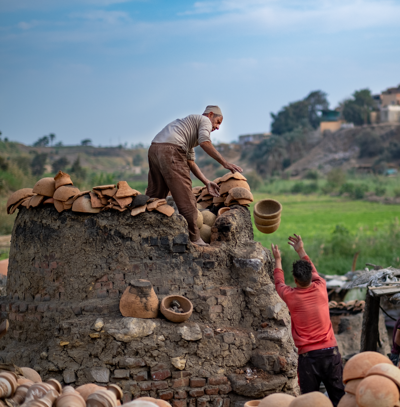 Homme debout sur une pile de terre, penché en avant et jetant un vase en poterie vers un homme debout, les bras levés, se trouvant en dessous de lui