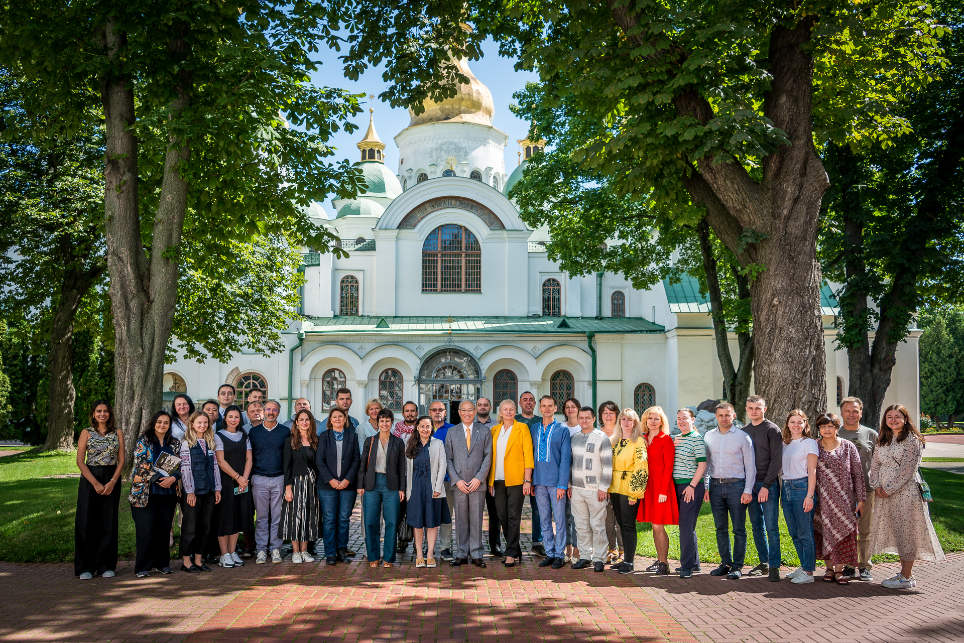 Group picture of 38 people standing and smiling between 2 tall trees. A white, green and golden religious building stands in the background.