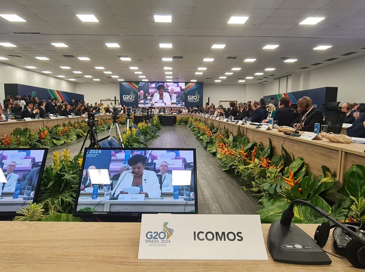 Room with tables placed in a rectangle and people sitting around them. A screen is placed on each wall of the room, it displays footage of a woman giving a speech.