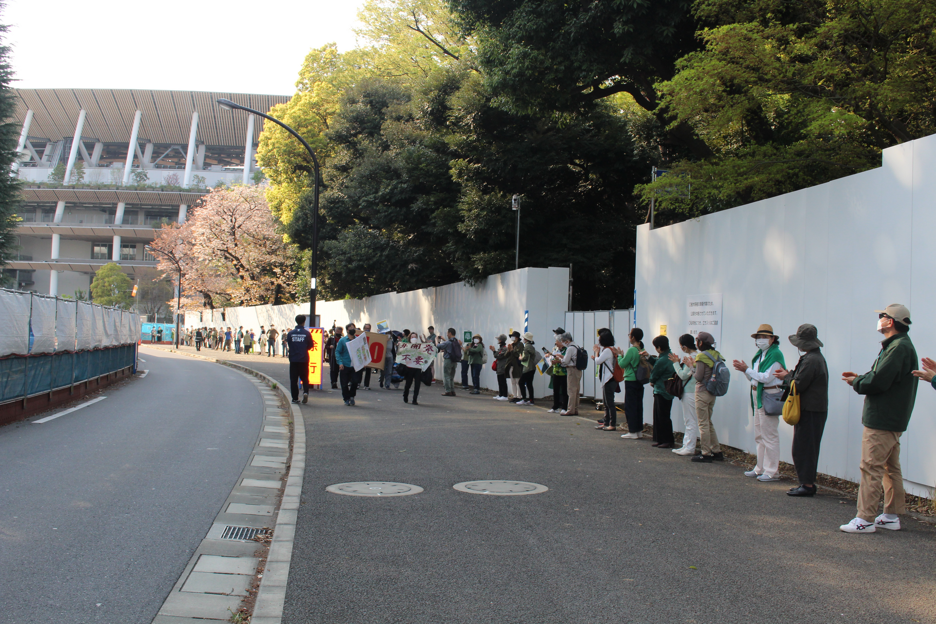 People standing in line and applauding in front of a white wall, behind which tall trees are peaking. A few protesters holding signs are walking along the line of people.