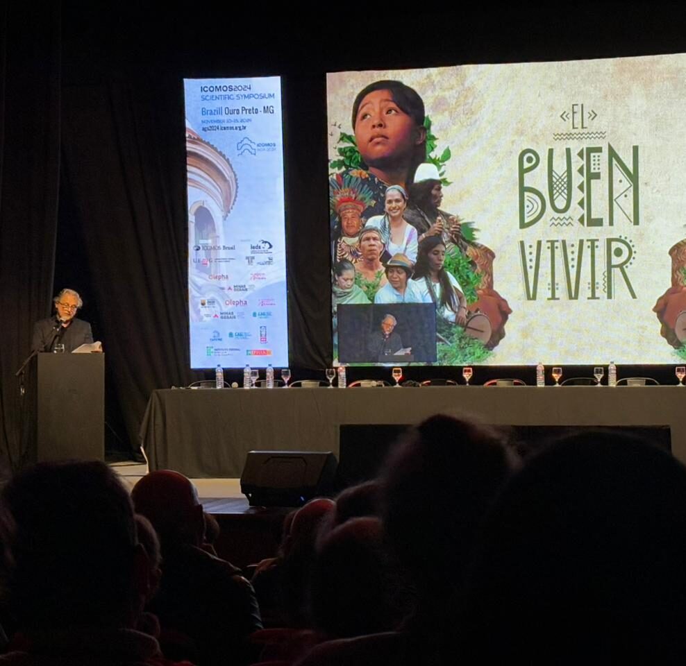 Man standing, facing a seated crowd, giving a presentation in front of a projected screen showing the words "el buen vivir"