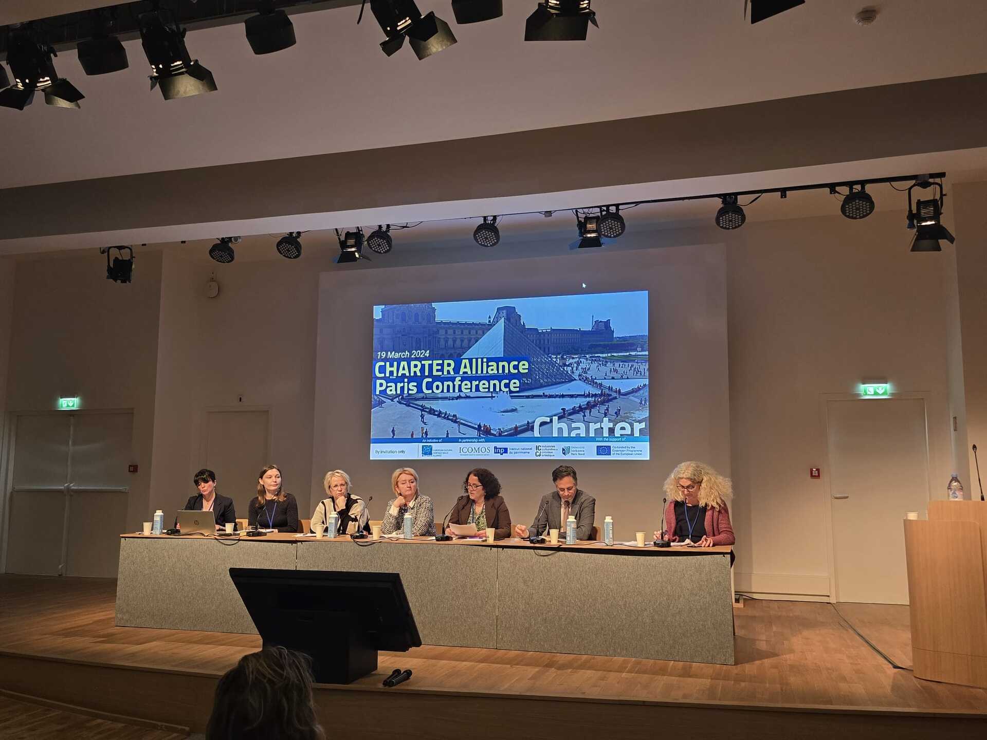 A group of 6 women and 1 man sitting behind a grey panel in front of a poster that says: CHARTER Alliance Paris Conference, 19 March 2024