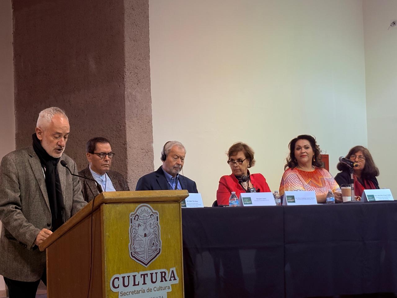 Picture showing the chairmanship of the 1st ICOMOS Scientific Symposium of the Americas : 5 people are sitting next to each other behind table, while 1 person is standing behind a reading desk and reading from a paper