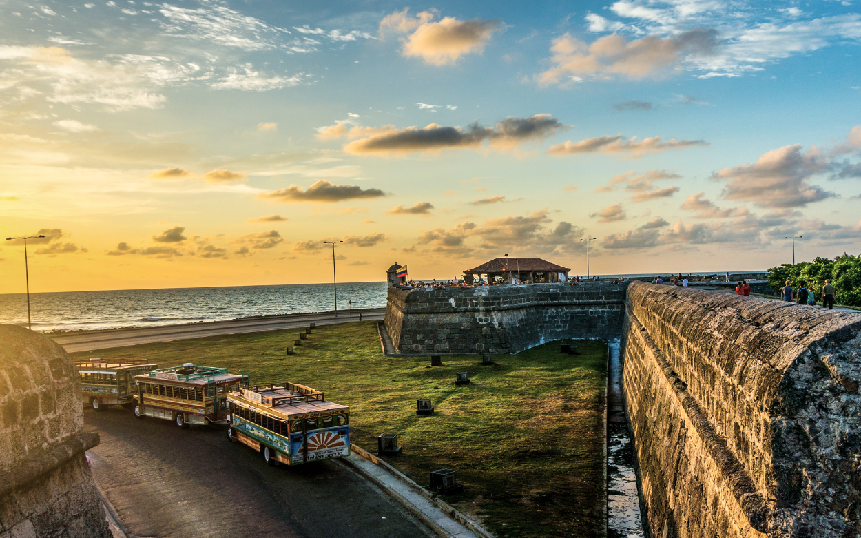 Picture of the Baluarte de Santo Domingo, a 17th century bastion located in Cartagena de Indias, in Colombia. The picture is taken from the walls, which overlook the sea, at sunset time. We can see parts of the fortifications and, in the background, the crowded Cafe del Mar.