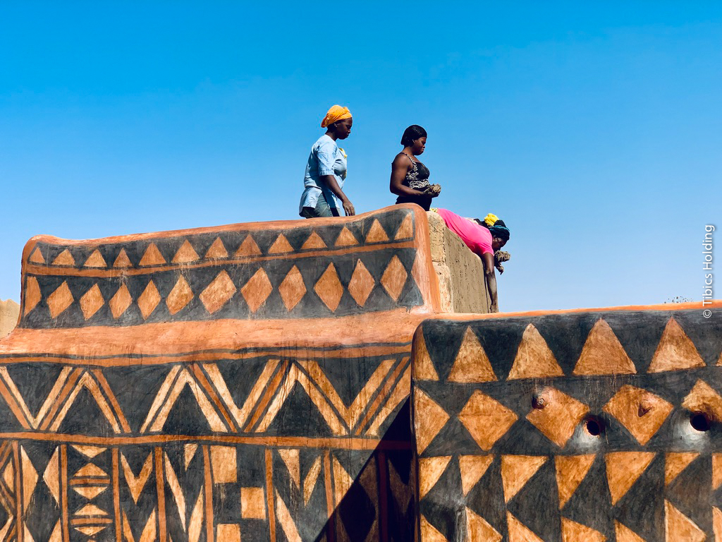 3 women from Burkina Faso are seen on the roof of a traditional building made of earthen architectural heritage which has been hand-painted and features black geometric shapes. One woman is bent over the roof and appears to be plastering a bare wall with earthen materials, while the other 2 women are watching her.
