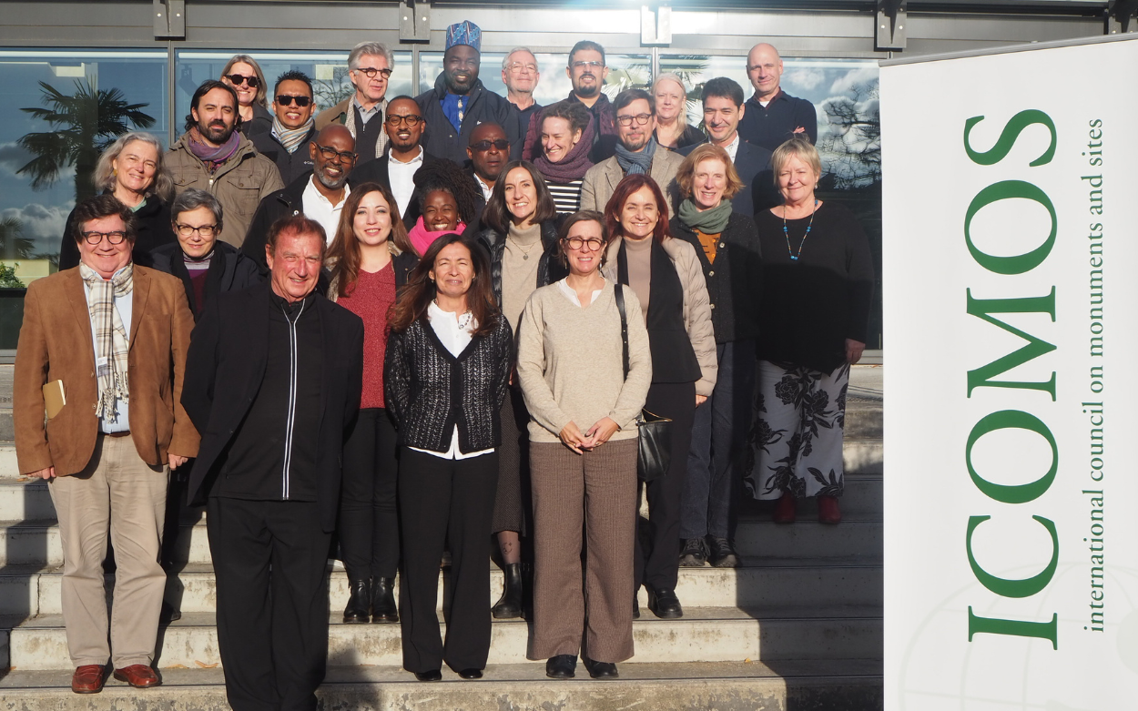 Picture showing the ICOMOS 2025-2026 Panel members posing outside on steps alongslide a roll-up featuring ICOMOS' logo